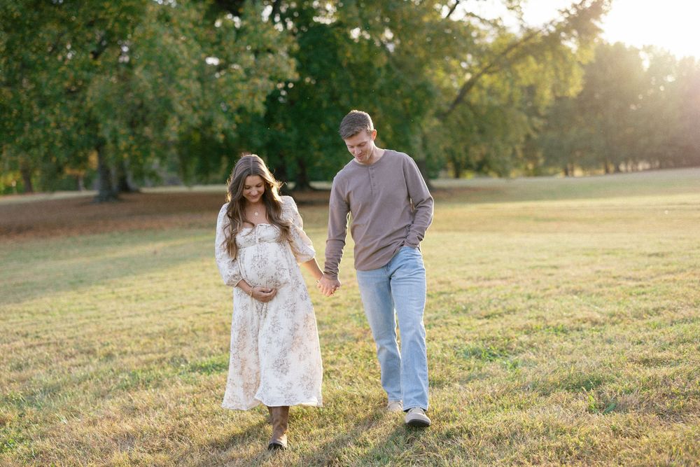 A couple walks hand in hand through a sunlit grassy field during golden hour at sunset.