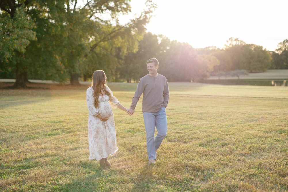 A couple walks hand in hand through a sunlit meadow during golden hour as sunlight streams through the trees.
