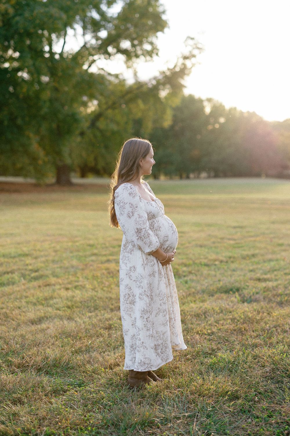 A pregnant woman in a white floral dress stands in a sunlit field at golden hour.