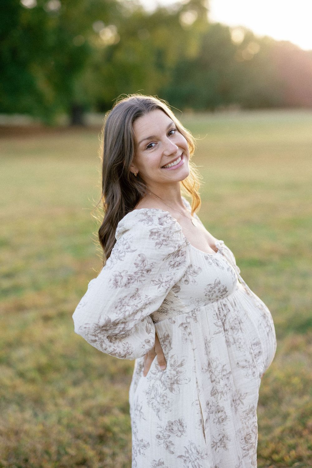 Woman in white lace dress poses outdoors at sunset in a grassy field during golden hour photo session.