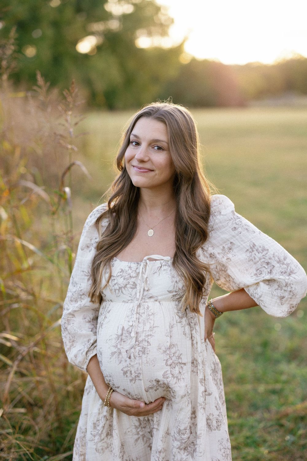 A woman in a flowing white floral dress poses in a sunlit field during golden hour.