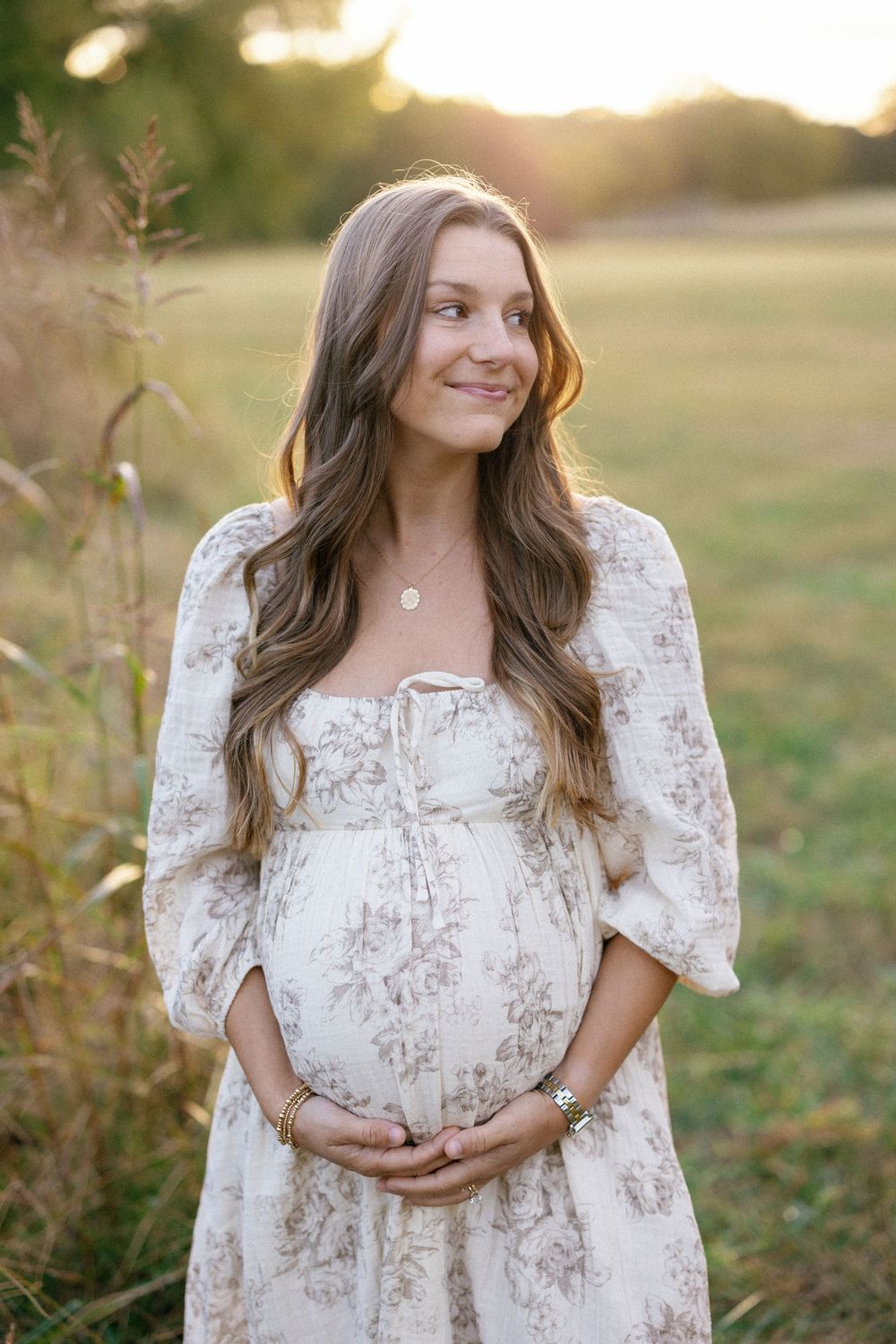 A woman in a white floral dress poses outdoors during golden hour sunset in a grassy field.