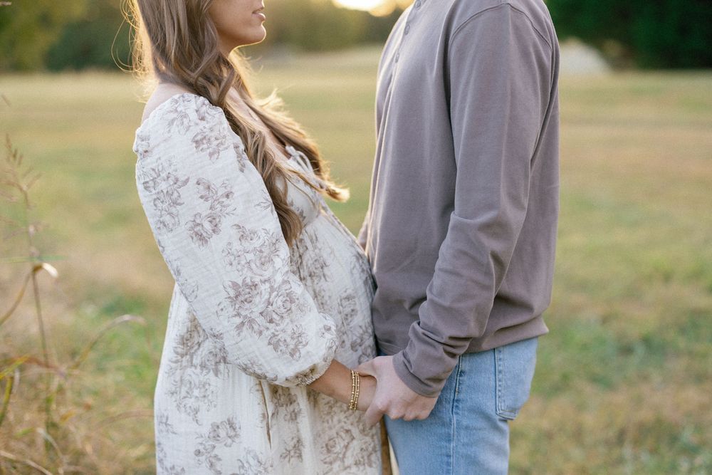 A couple stands together in a sunlit field wearing white lace and gray casual attire during a romantic photoshoot.