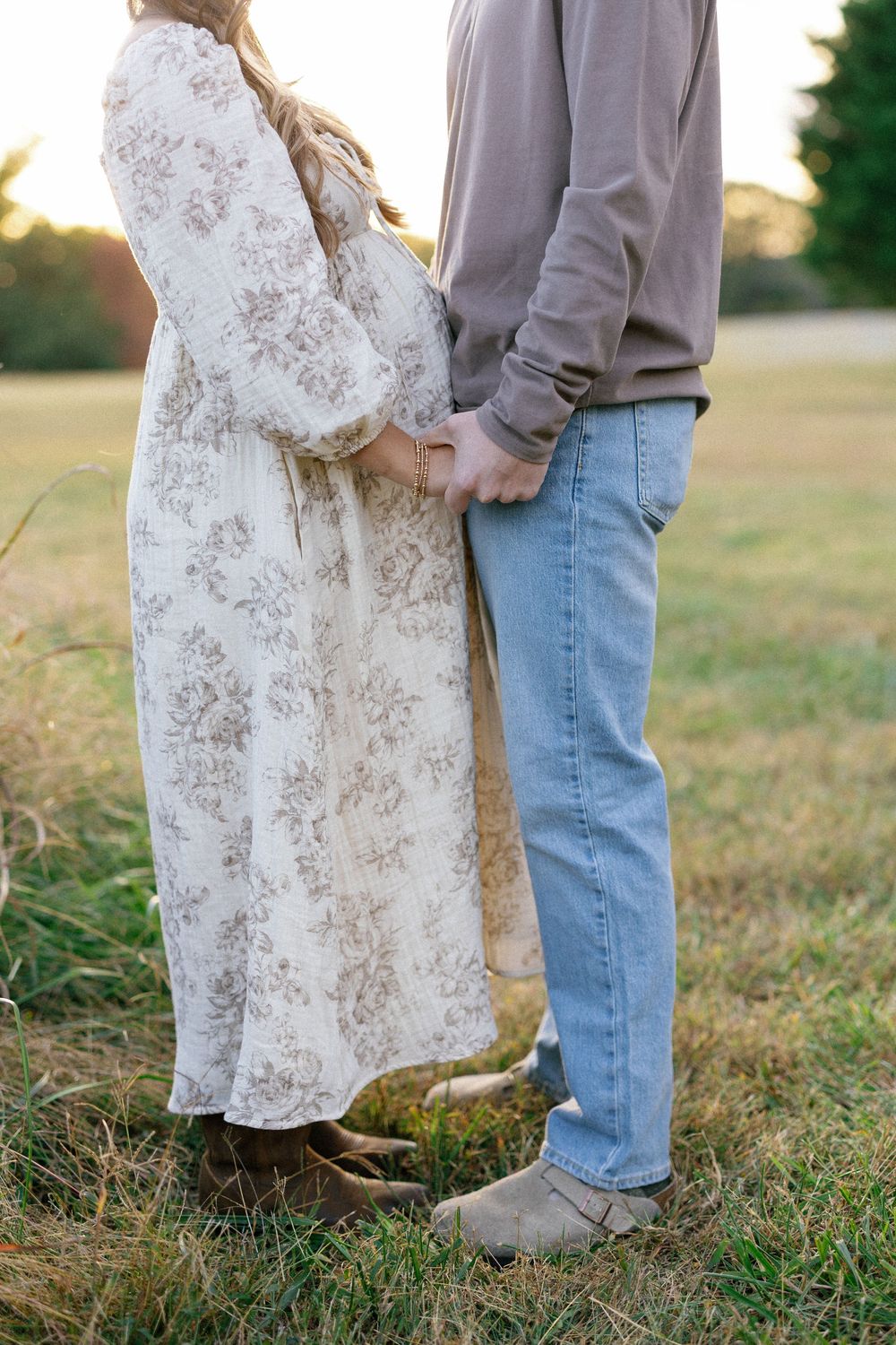 Couple in casual attire embracing in a grassy field during golden hour sunset.