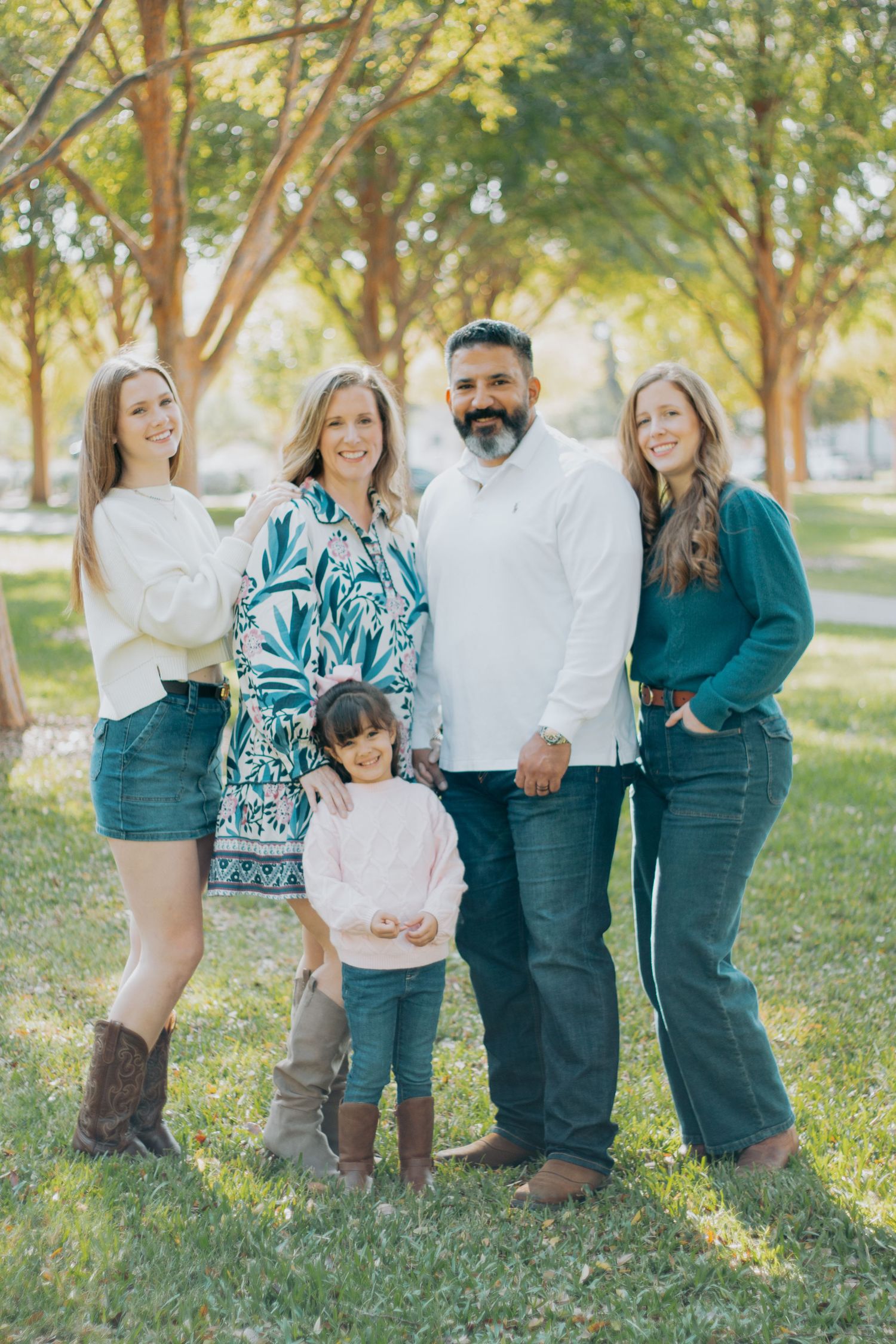 A group poses together outdoors in casual attire against a backdrop of sunlit trees on a grassy lawn.