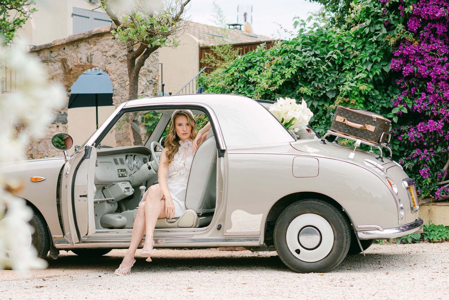 A vintage silver luxury car is photographed from multiple angles in a garden setting with blooming purple flowers.