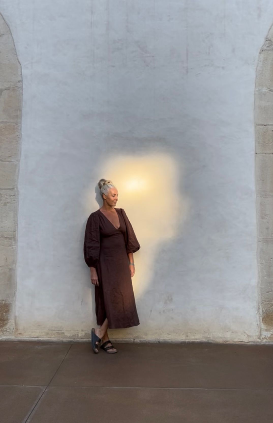 A woman standing alone against a pale textured wall, wearing a long dress and gazing into soft natural light.