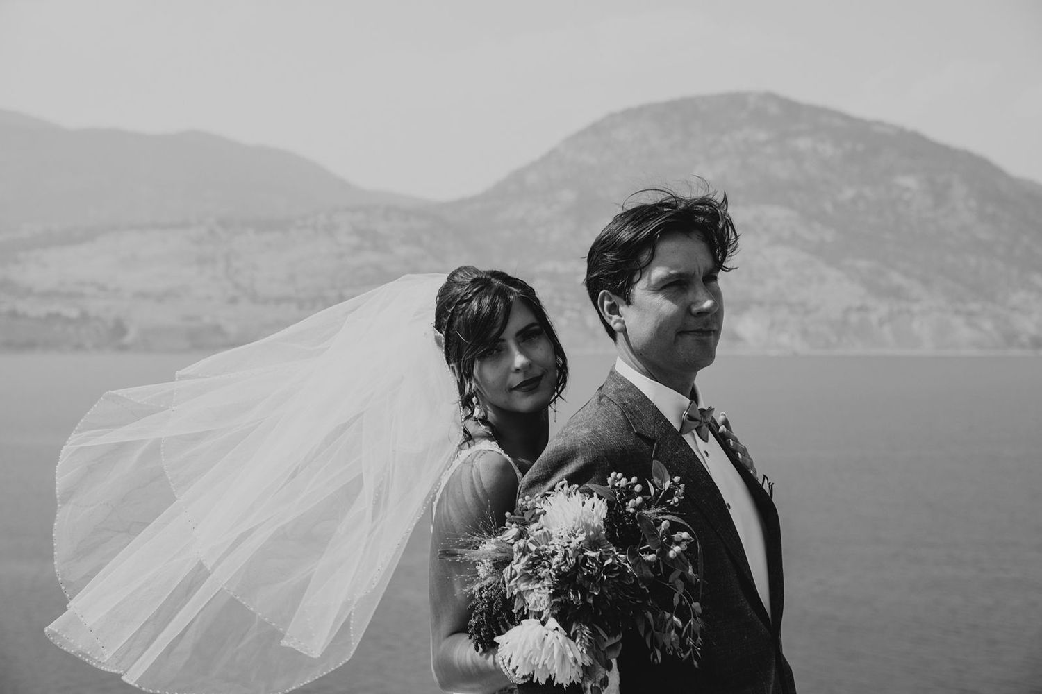 Black and white portrait of bride and groom back-to-back, veil flowing, with lake and mountains behind.