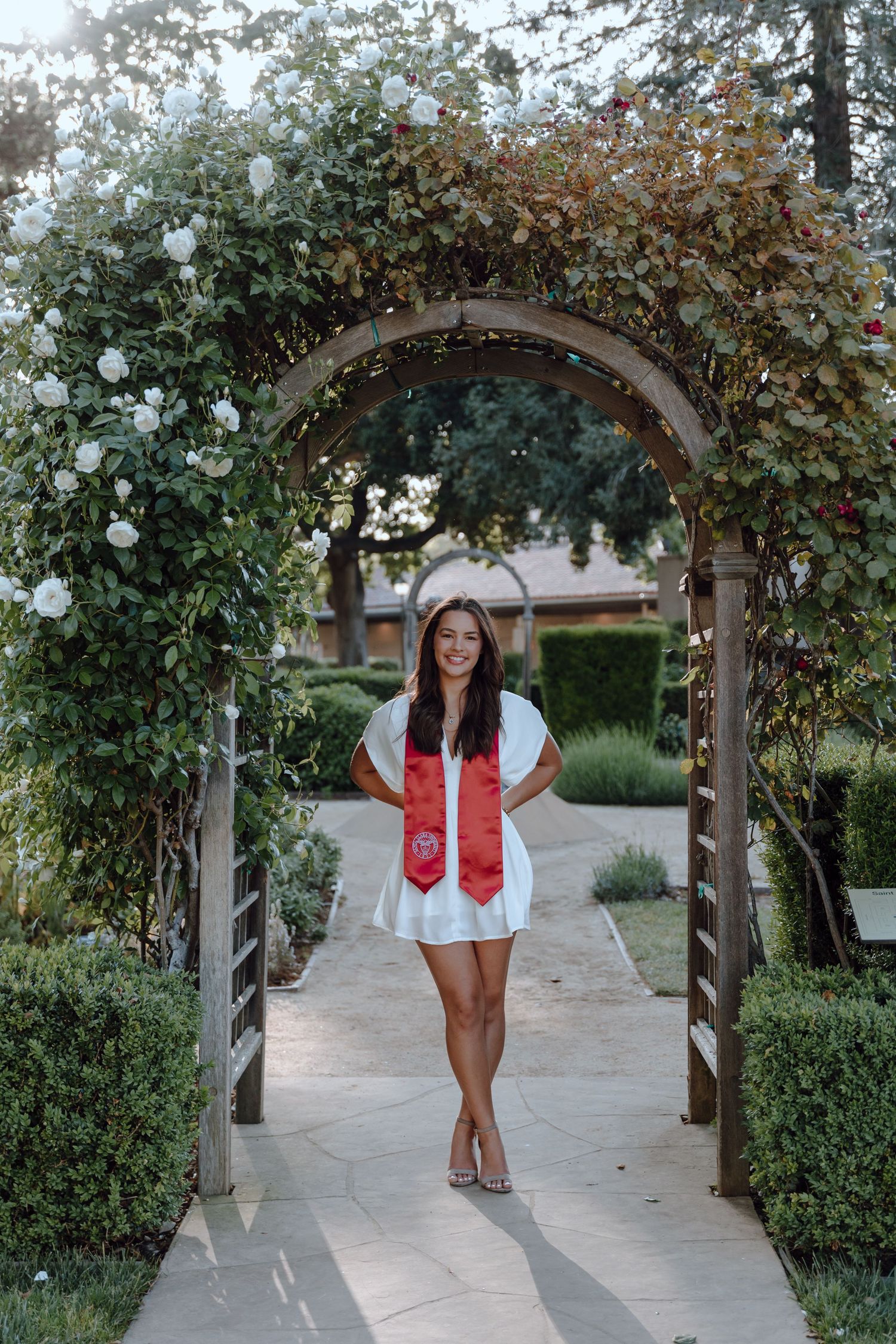 Santa Clara University graduate poses for her grad photoshoot by standing under an archway with flowers. By Bay Area Grad Photographer.
