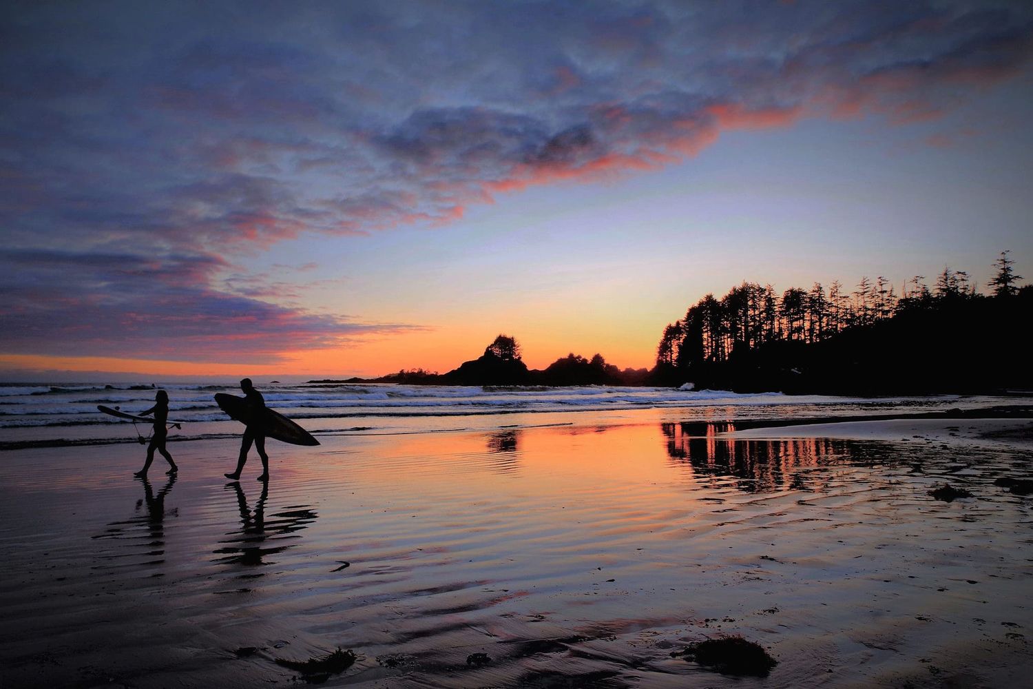 couple walks along the beach in Tofino with surf boards at sunset
