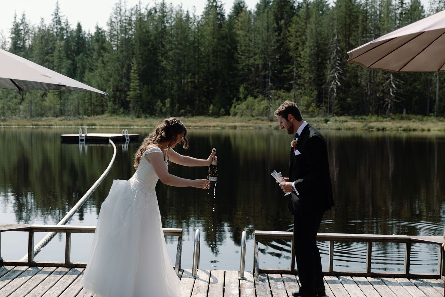 Bride and groom celebrate on a lakeside dock, opening champagne at their outdoor wedding.