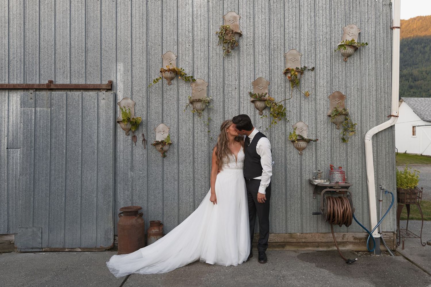 Bride and groom share a tender kiss against a rustic corrugated metal wall with floral decor.