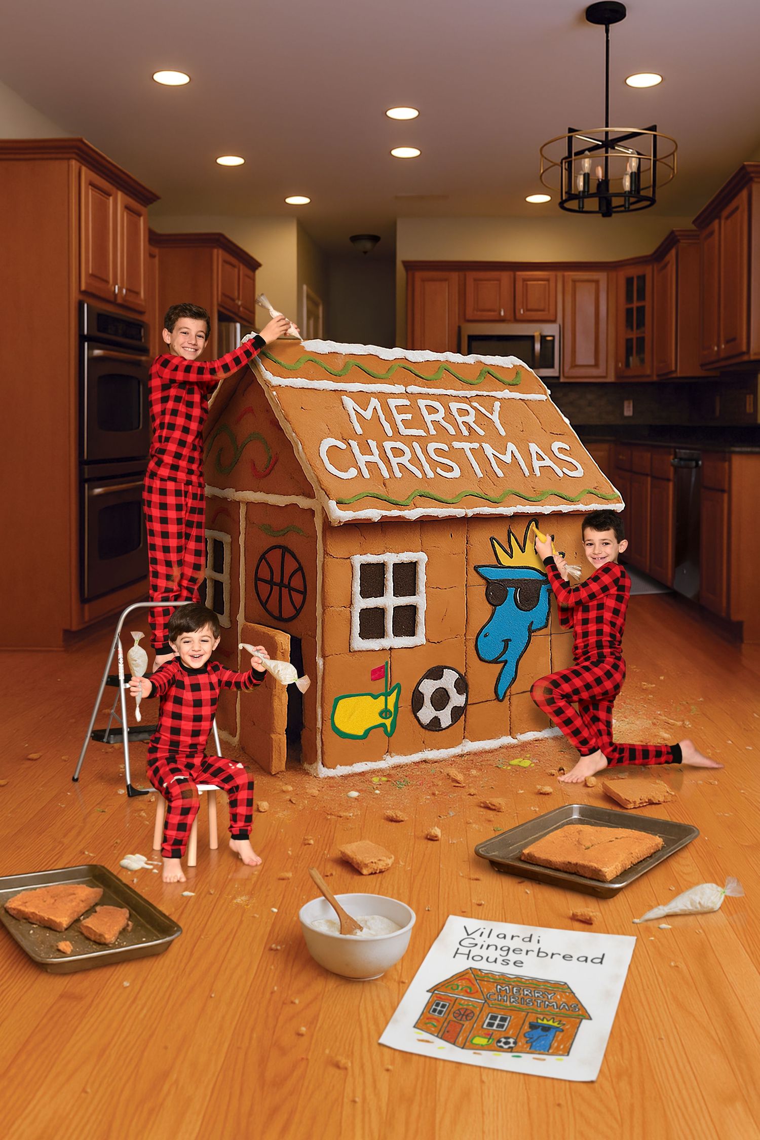 Children in matching plaid pajamas decorating giant gingerbread house in kitchen.