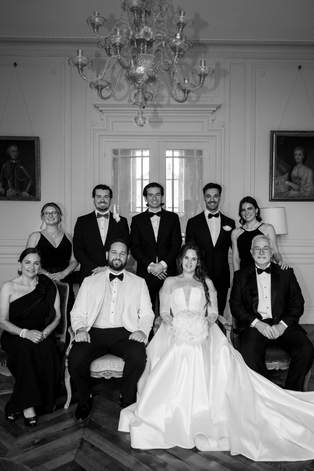 Formal black and white group portrait in an elegant room with chandeliers and ornate decor.