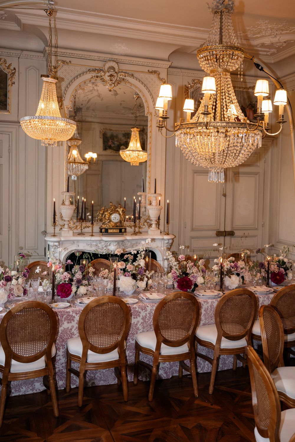 Elegant dining room with crystal chandeliers, ornate fireplace and table set for formal dinner with floral arrangements.