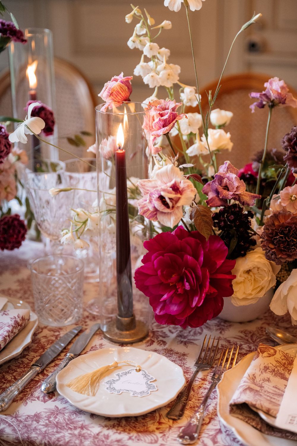 Romantic dining table with deep burgundy roses, crystal stemware, and candlelight on a vintage-style patterned tablecloth.