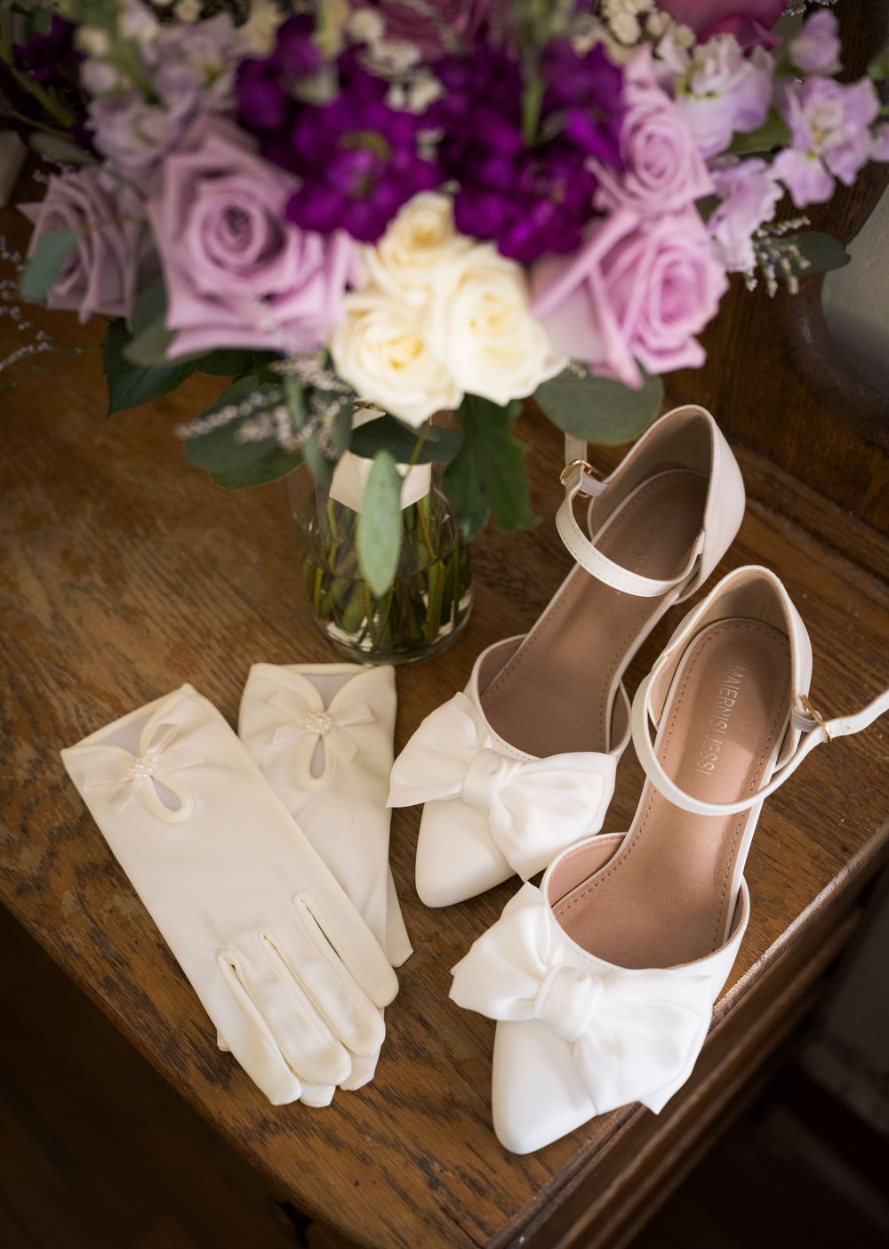 White bridal shoes and gloves arranged on wooden surface with pink and purple floral bouquet in background.