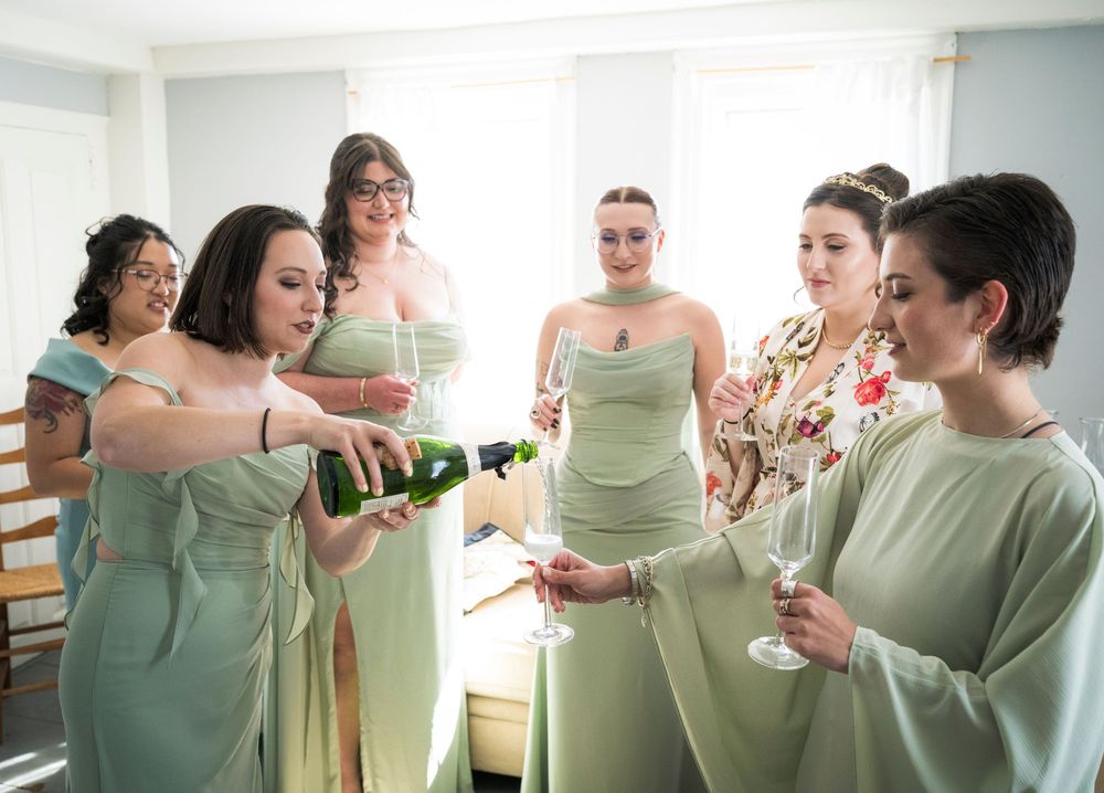 A group in mint green dresses pour champagne into glasses while getting ready in a bright room.