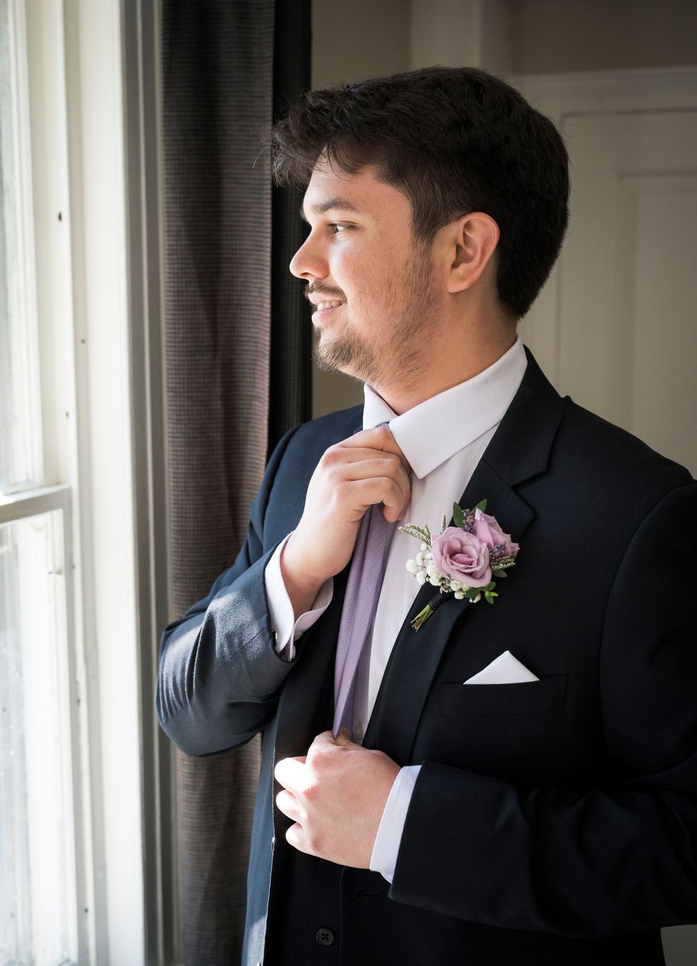 Groom in black suit with pink boutonniere adjusts his tie while looking in a window reflection.