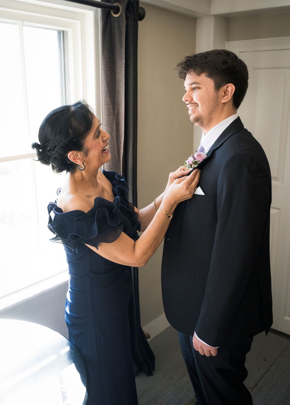 A woman in a navy dress adjusts a man's black suit jacket while standing near a bright window.