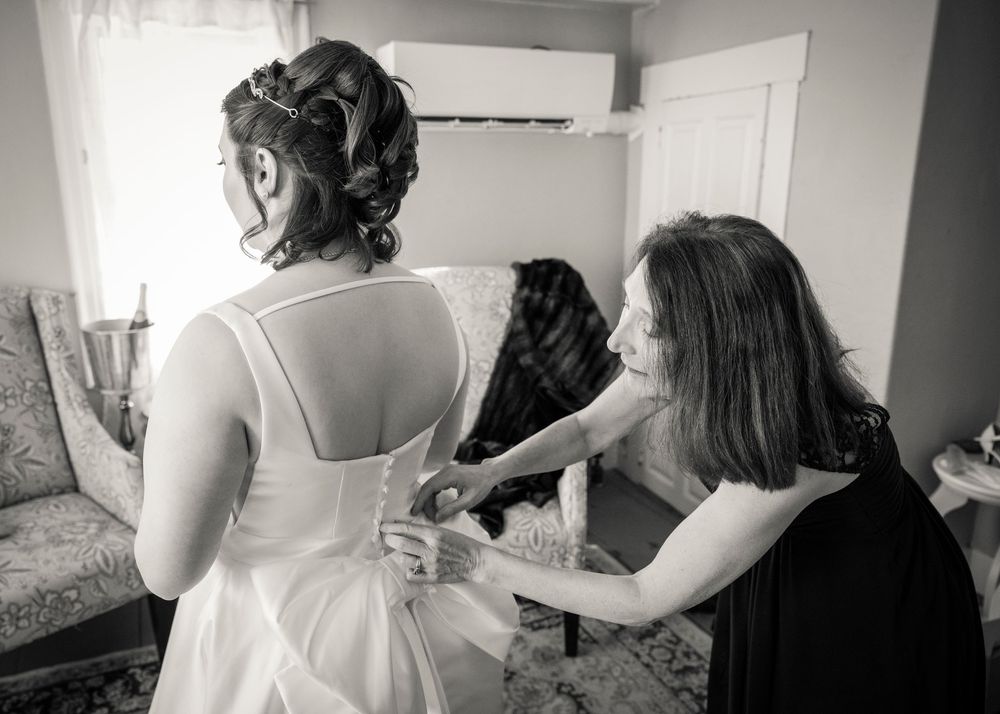 A bride gets help with final adjustments to her wedding dress while getting ready in a bright room.