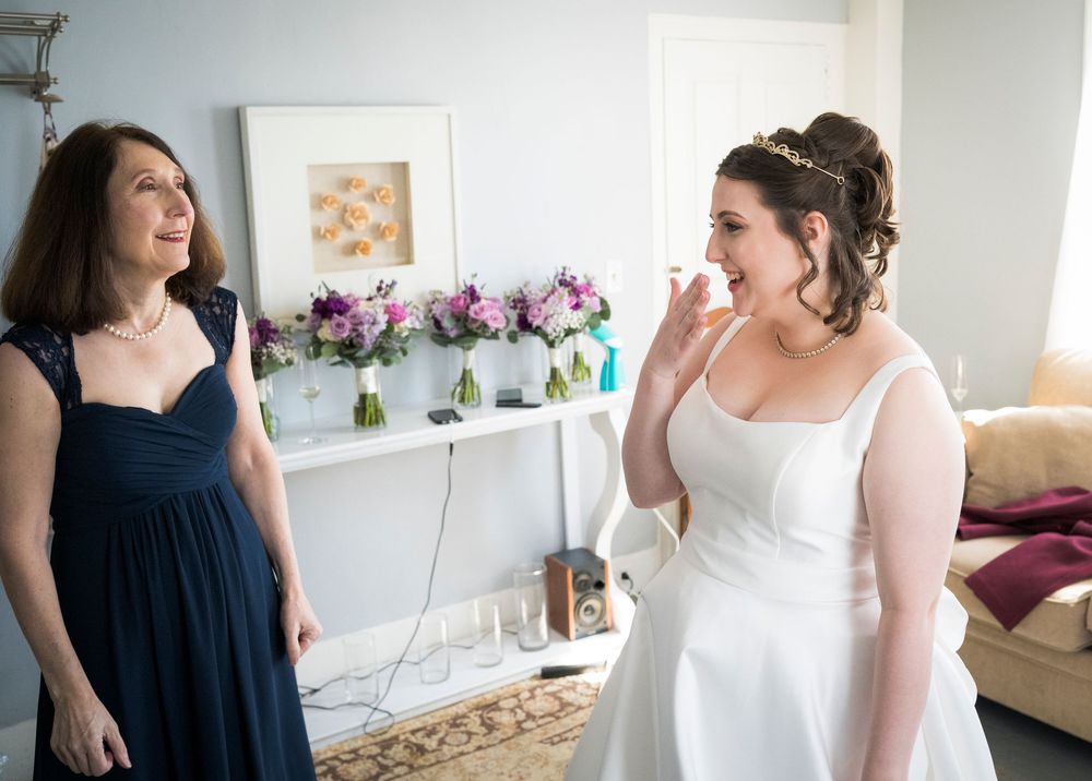 Two women in elegant dresses share a joyful moment while getting ready in a room decorated with purple flowers.
