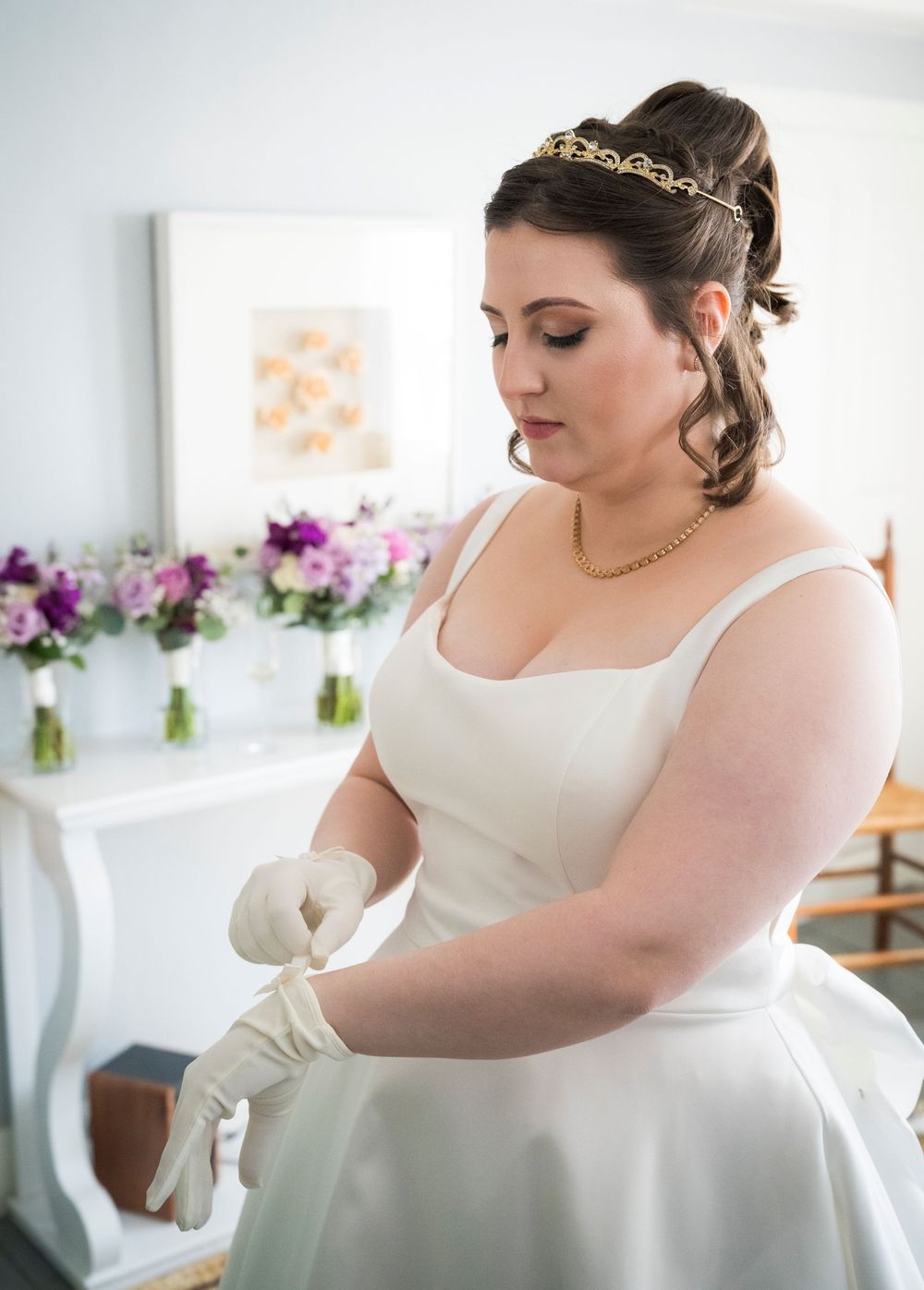 A bride in a white dress adjusts her gown while getting ready in a room decorated with purple flowers.