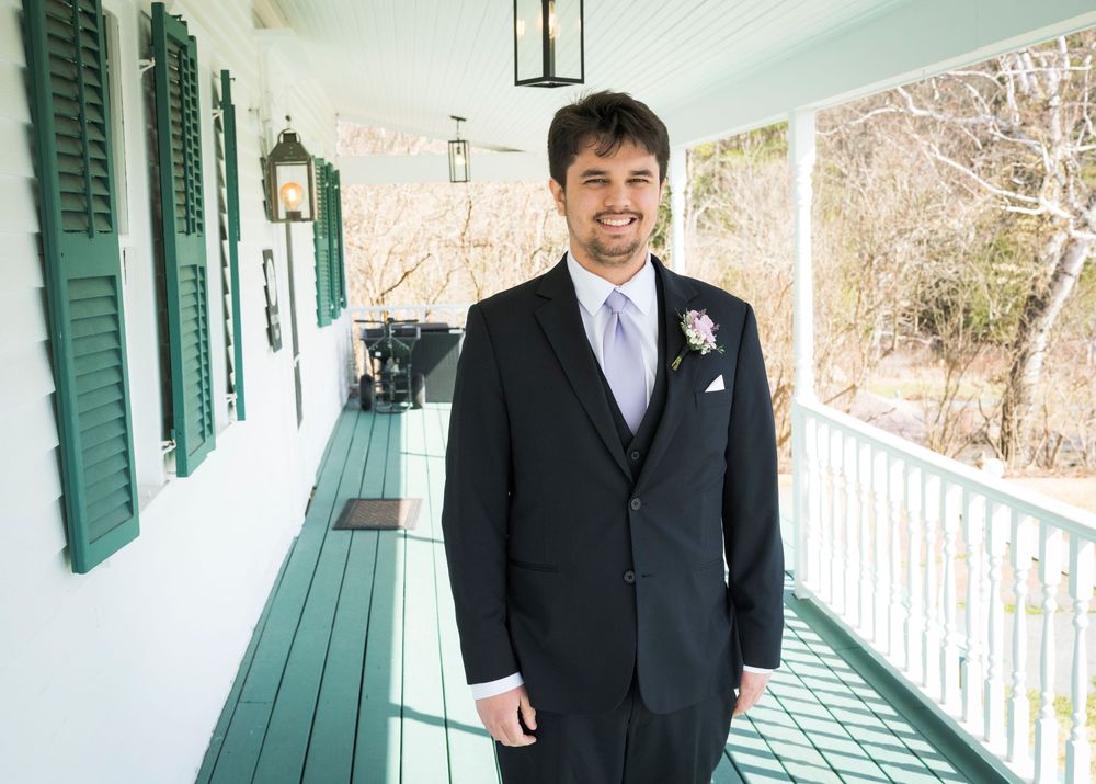 A groom in a black suit and purple tie stands smiling on a white porch with green shutters and benches.