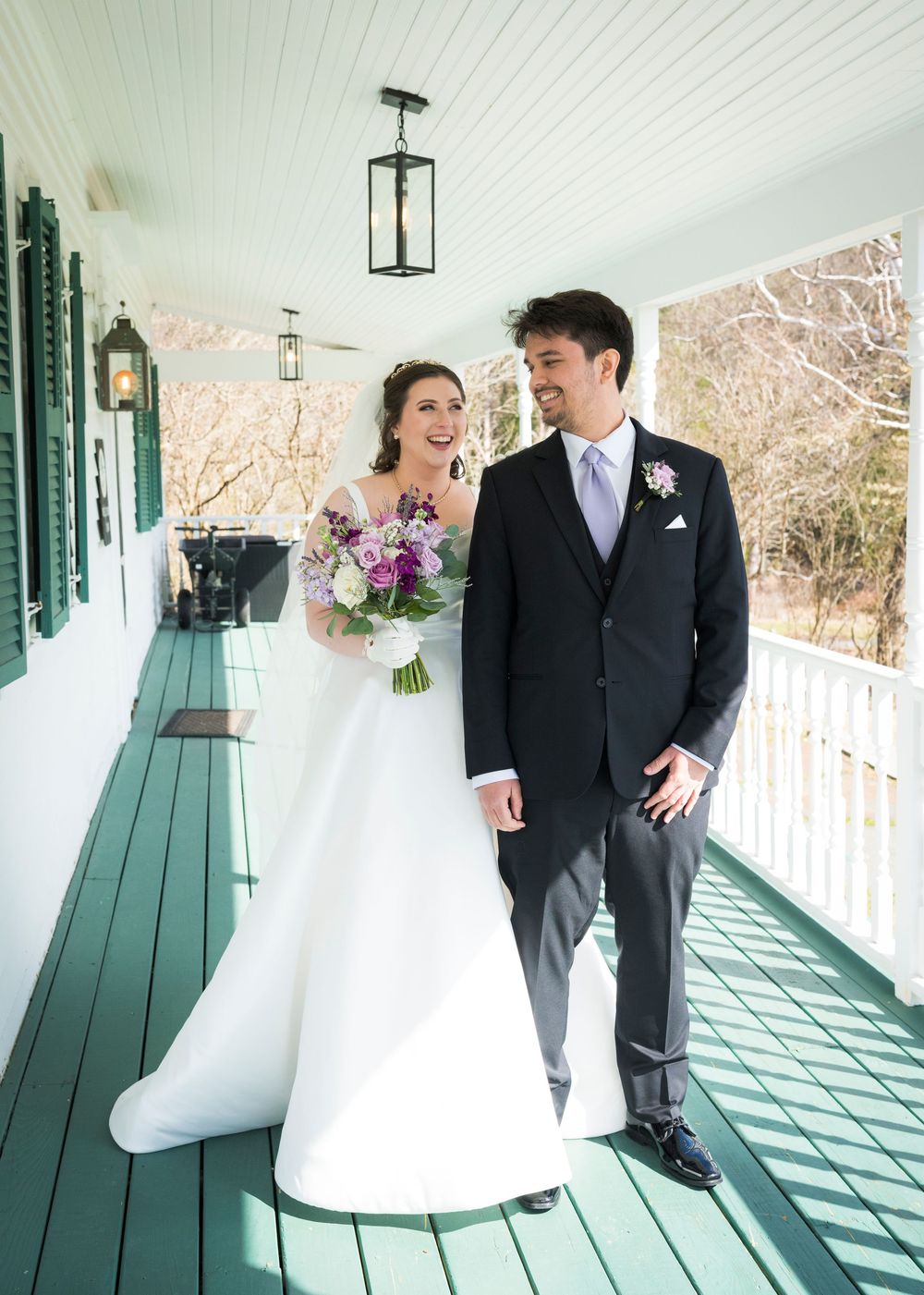 Elegant wedding couple poses on a white porch with green shutters and shadows creating beautiful patterns.