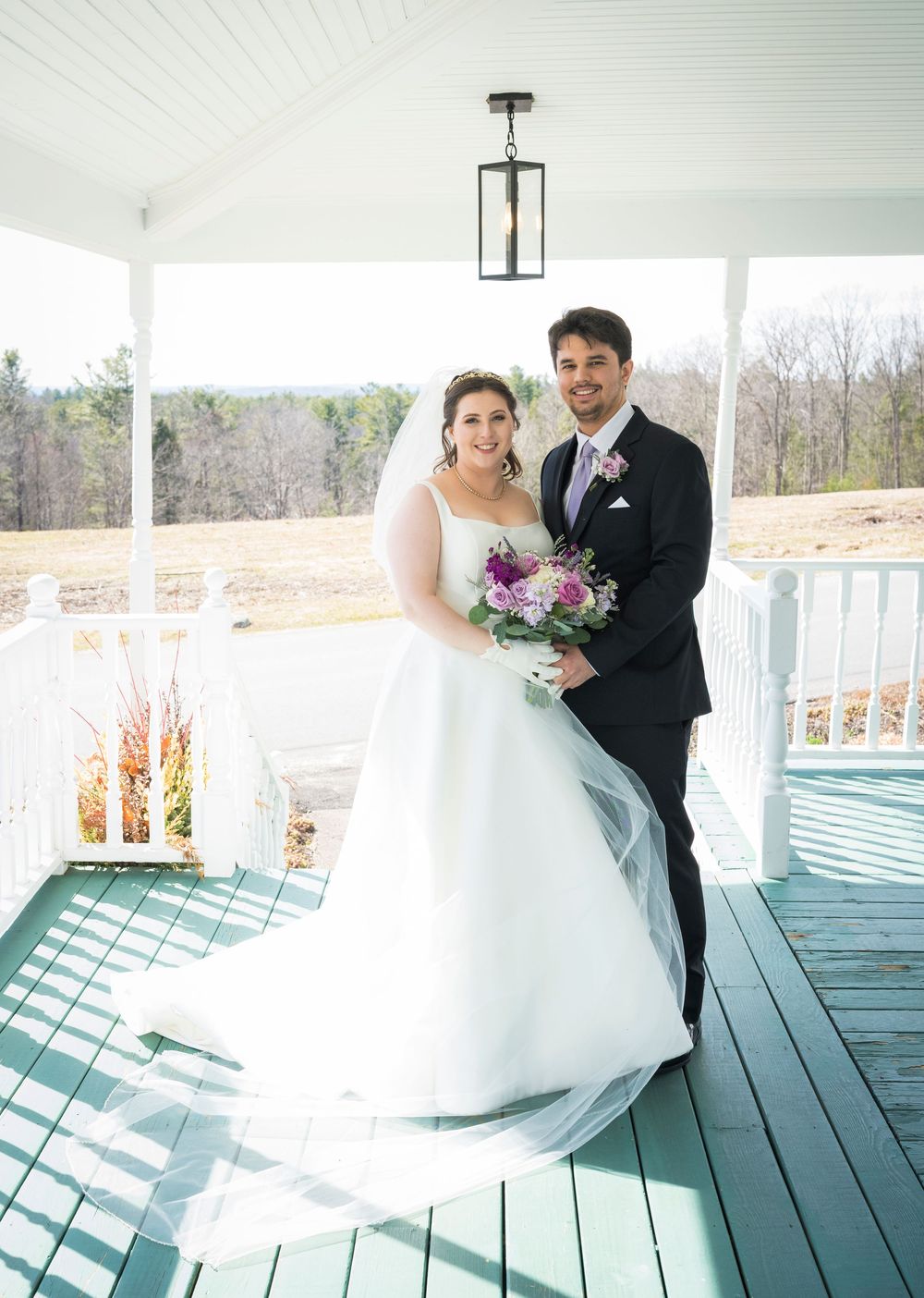 A wedding couple poses on a covered porch with white columns and a lantern pendant light overhead.