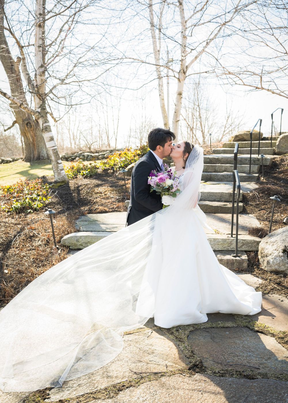 Wedding couple shares romantic moments on stone steps surrounded by bare winter trees.