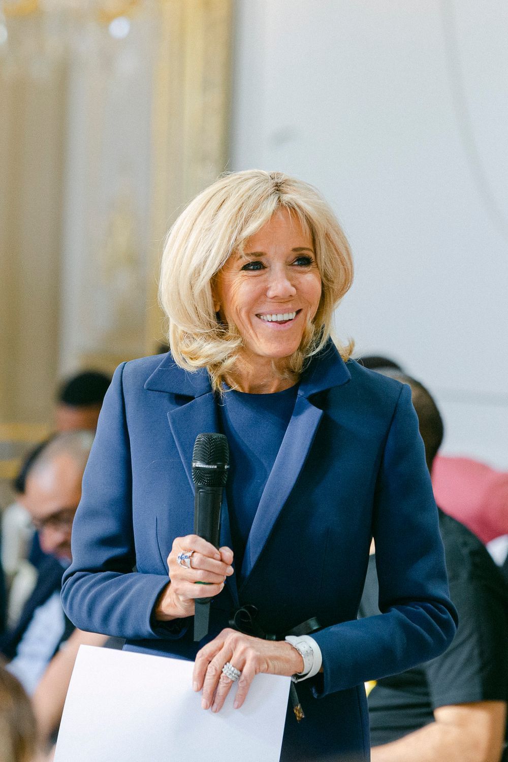 A person in a navy blue blazer speaks into a microphone while gesturing during a presentation.
