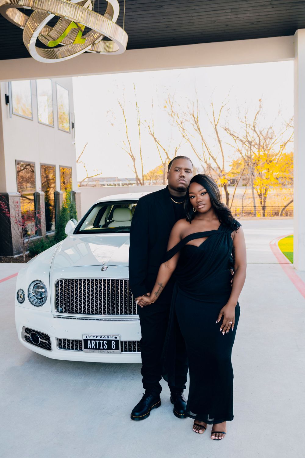 A couple poses together in front of a white Bentley luxury car at sunset.