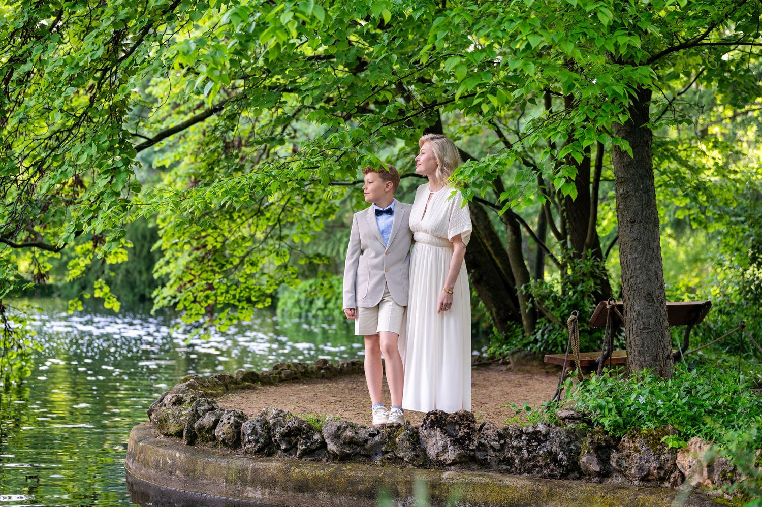 A couple in light summer clothing stands together on a wooded path surrounded by lush green foliage.