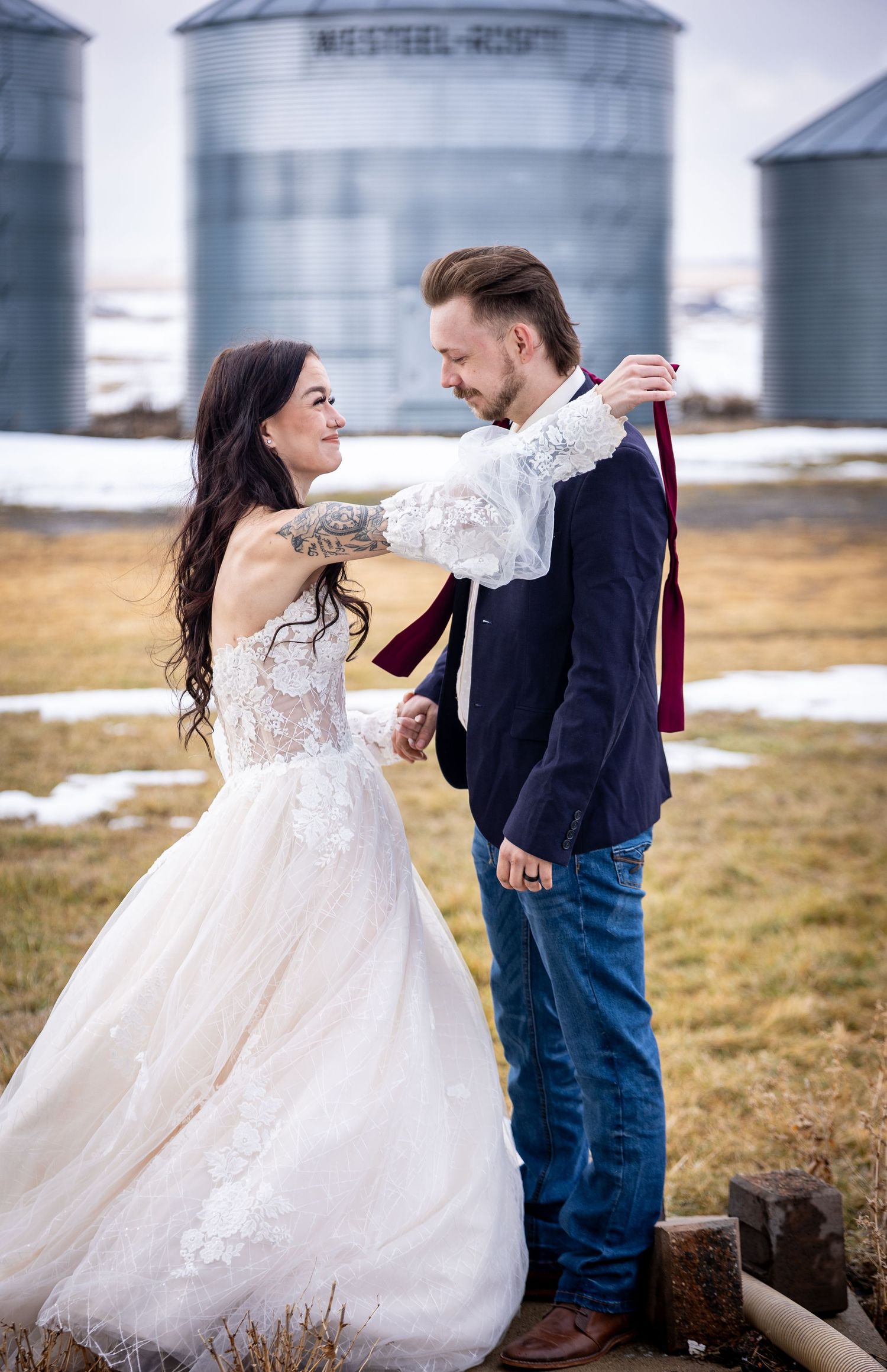A romantic couple dances playfully in a snowy field with grain silos in the background, the bride's dress flowing in the wind.