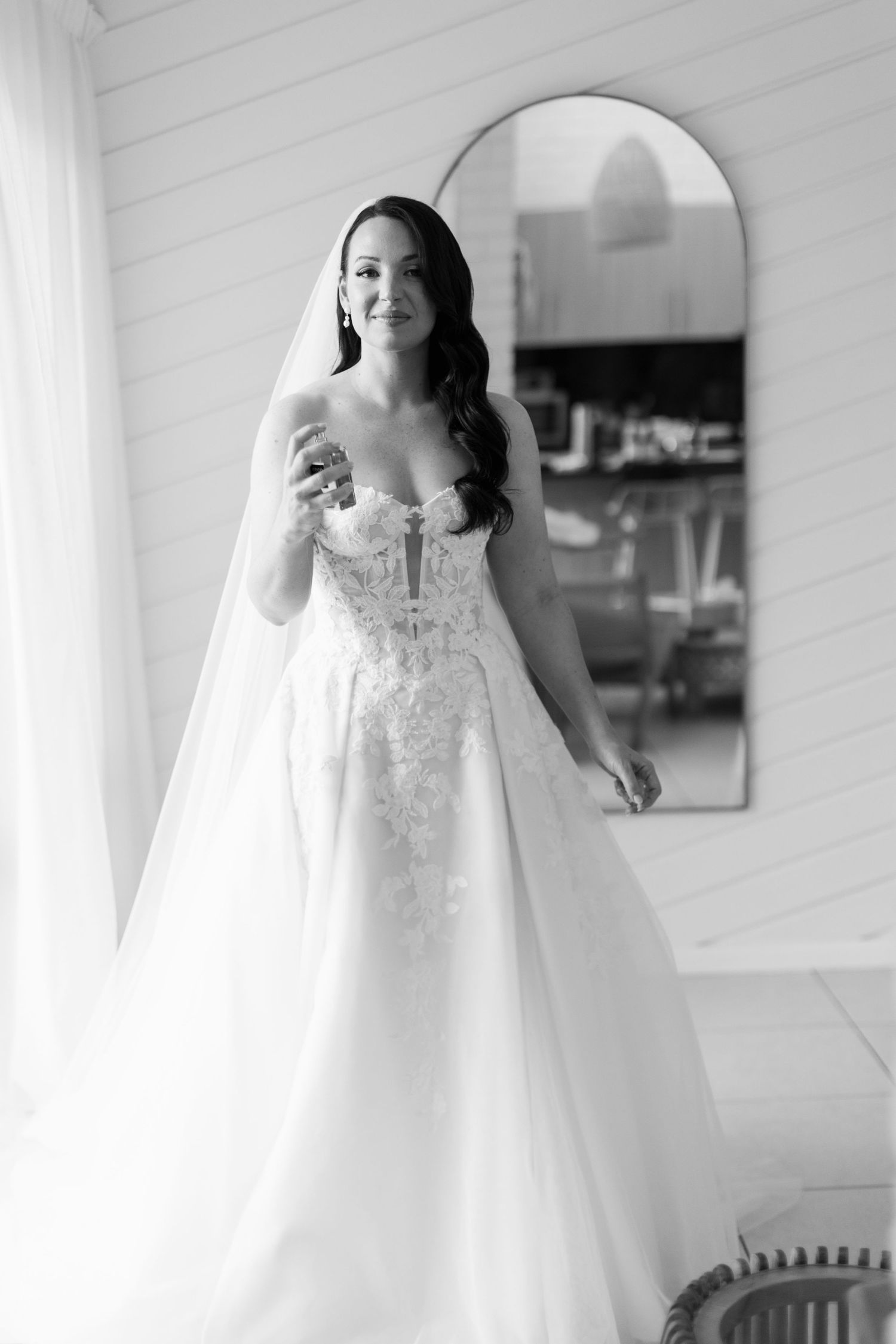 A bride in an elegant white lace wedding dress and veil twirls gracefully in a bright indoor setting.