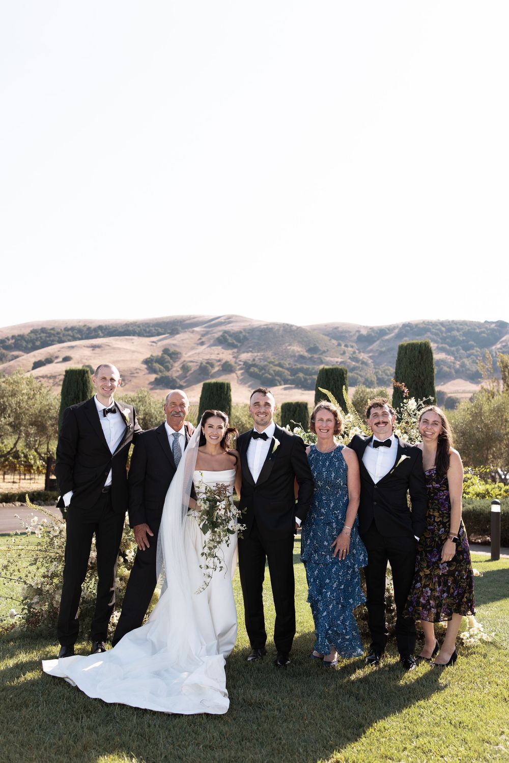 A wedding party poses together outdoors on a grassy lawn with rolling hills in the background.