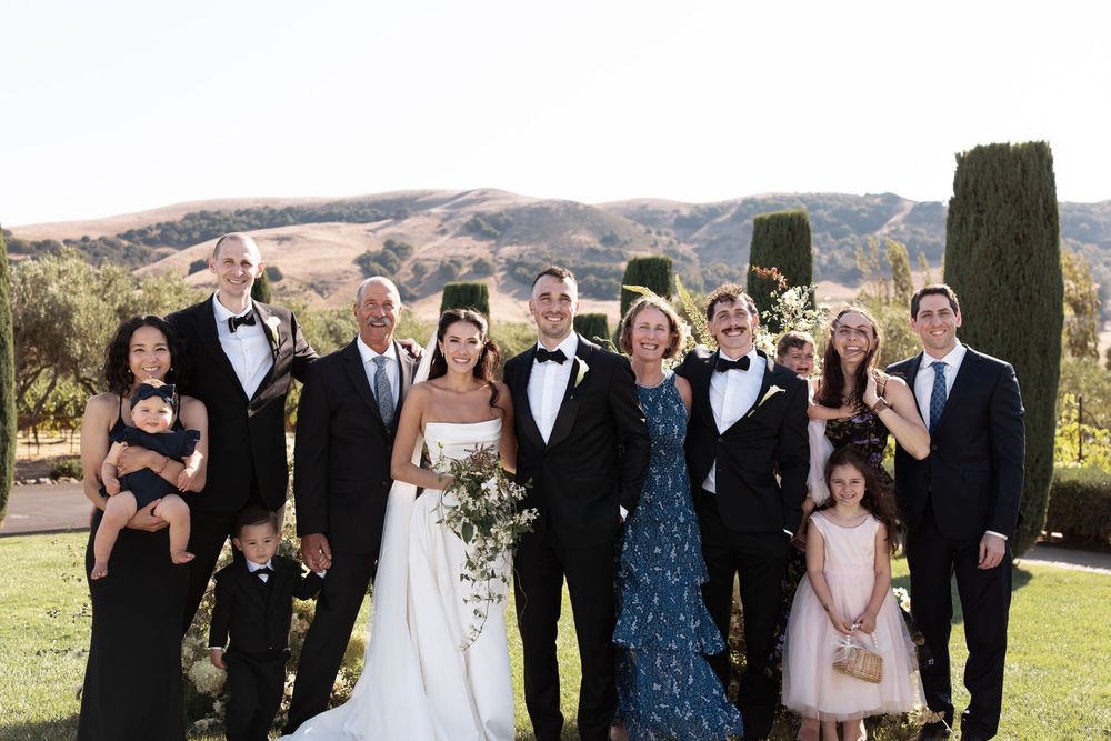 Wedding party poses together on a sunny day at an elegant outdoor venue with rolling hills in the background.