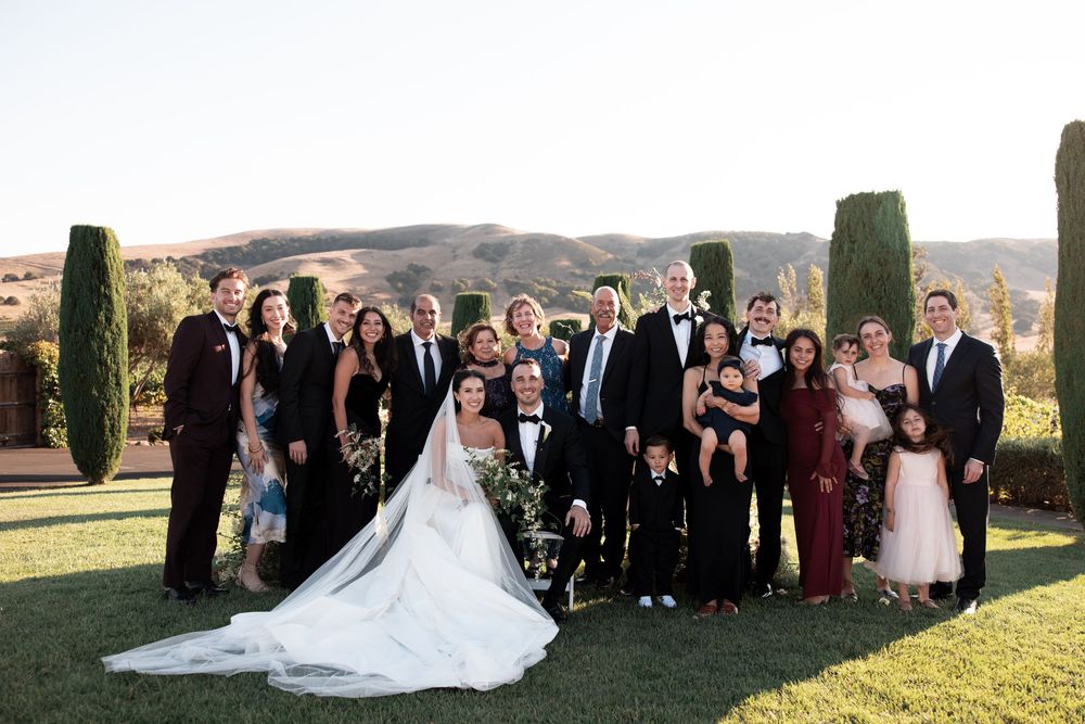 Wedding party gathers on a scenic hilltop during golden hour for formal group photos at an outdoor ceremony.