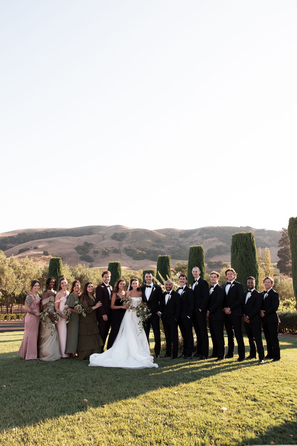Wedding party in formal attire poses outdoors on grassy hill with scenic mountain backdrop during autumn.