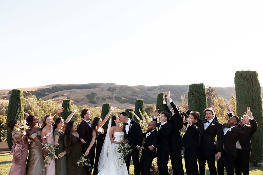 Wedding party celebrates outdoors with raised arms against backdrop of rolling hills and cypress trees at sunset.
