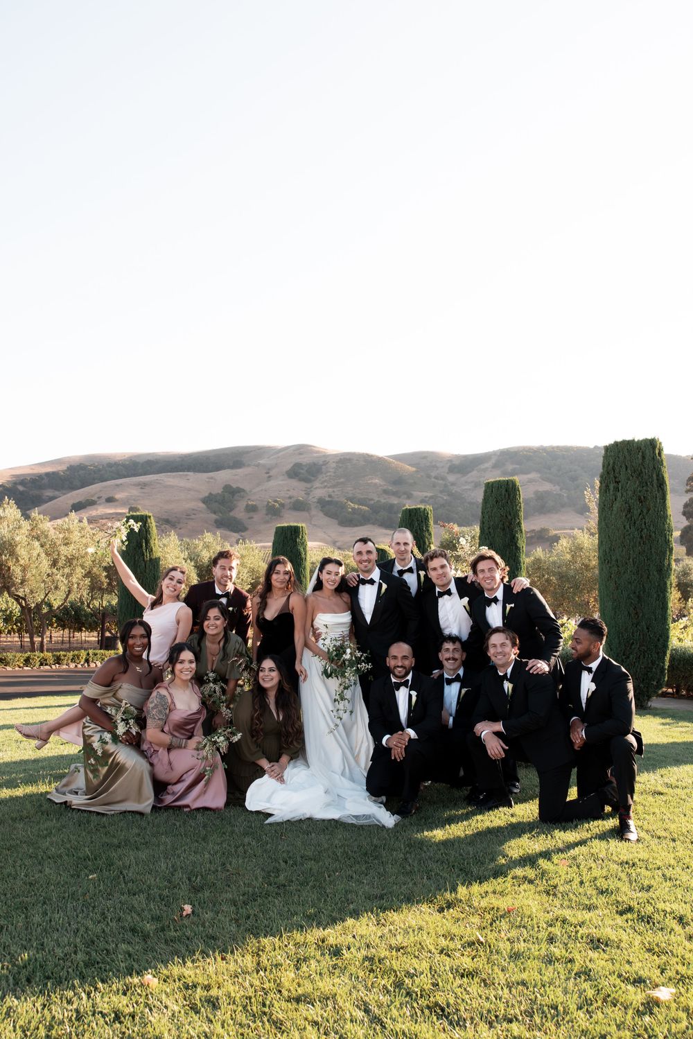 Wedding party in formal attire poses together on a grassy hillside during golden hour at an outdoor venue.