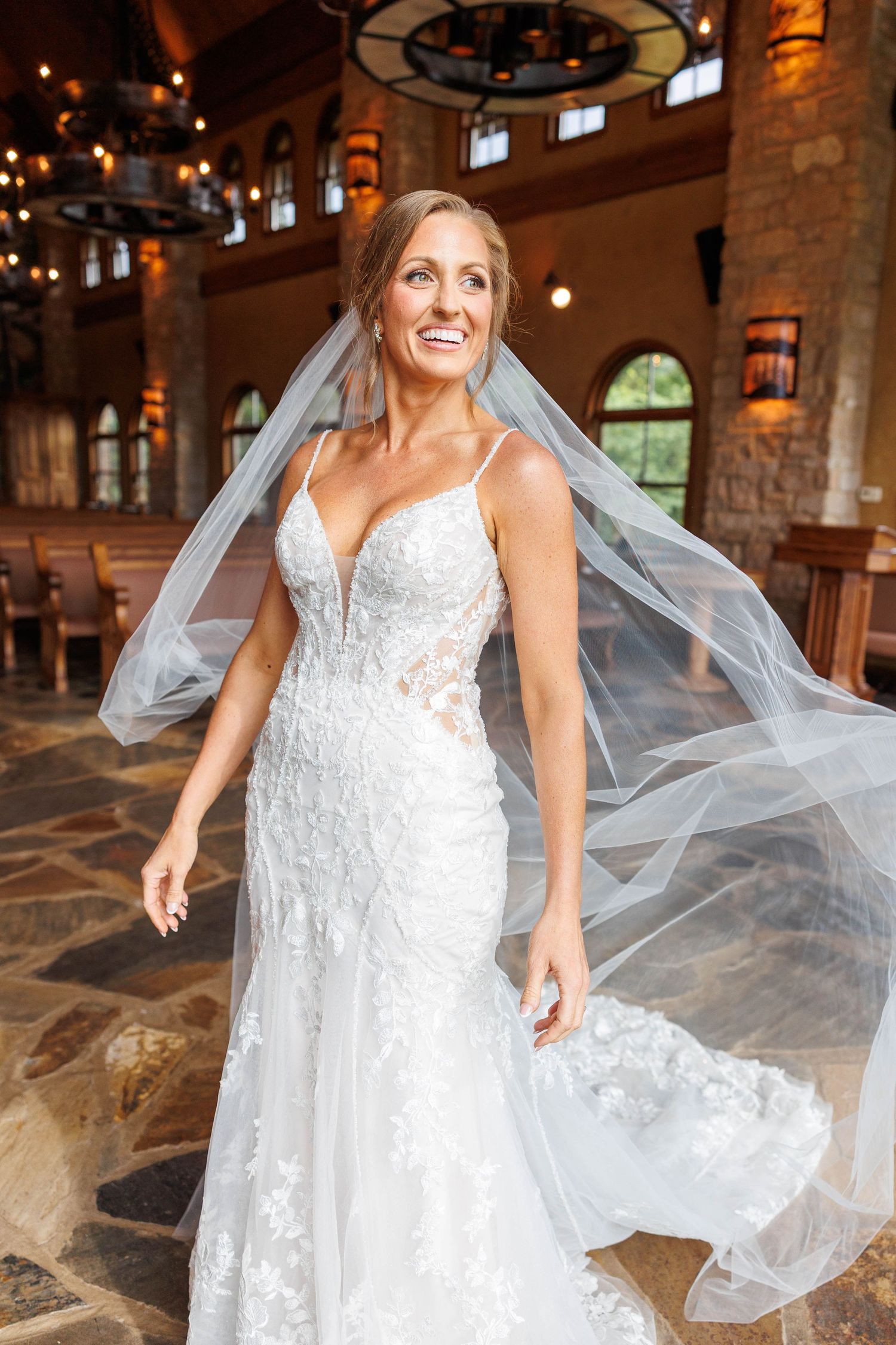 A bride in a white lace wedding gown with flowing veil twirls joyfully inside a rustic stone venue with warm lighting at Integrity Hills.