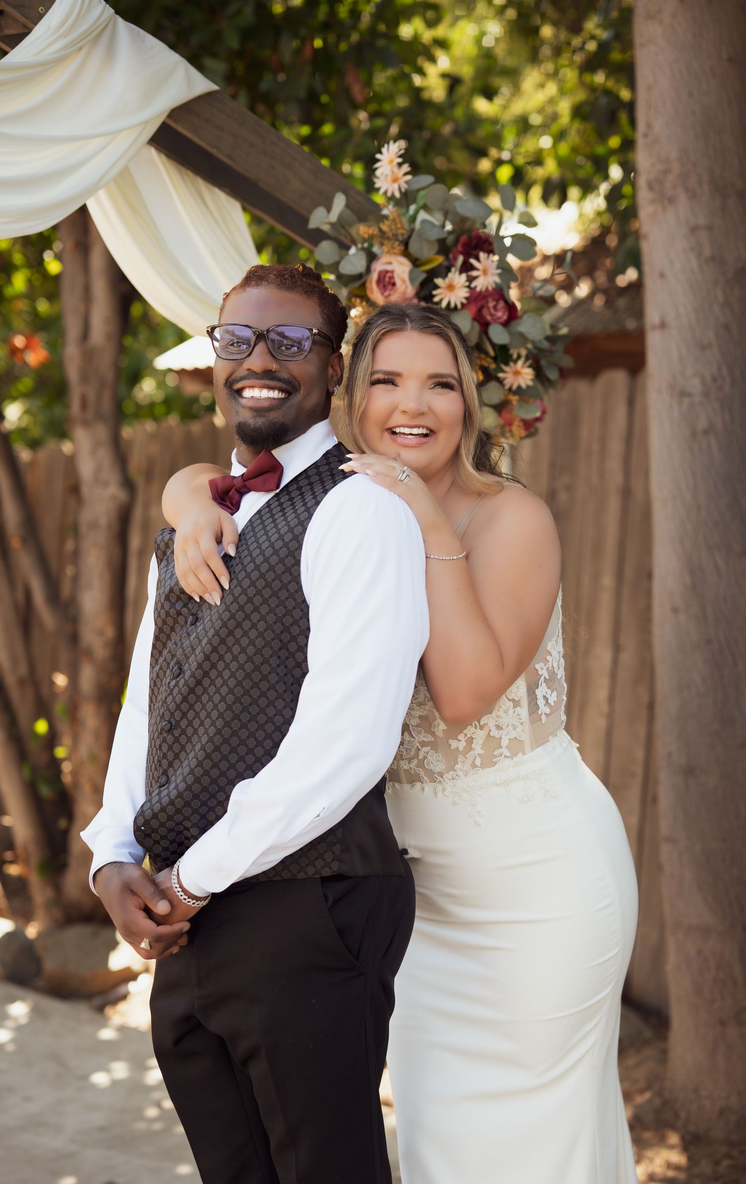 A joyful bride and groom share candid laughs during their outdoor wedding photo session under trees.