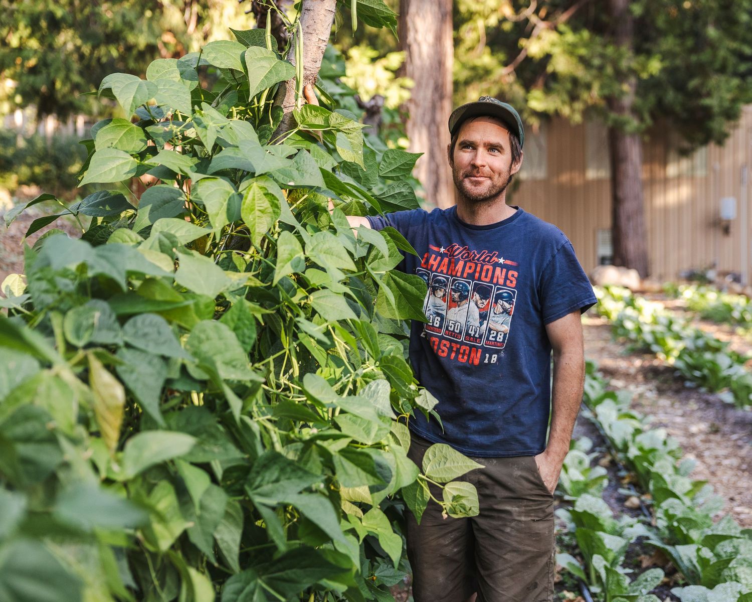 Person in blue t-shirt stands smiling in a lush garden with green plants and foliage in the background.