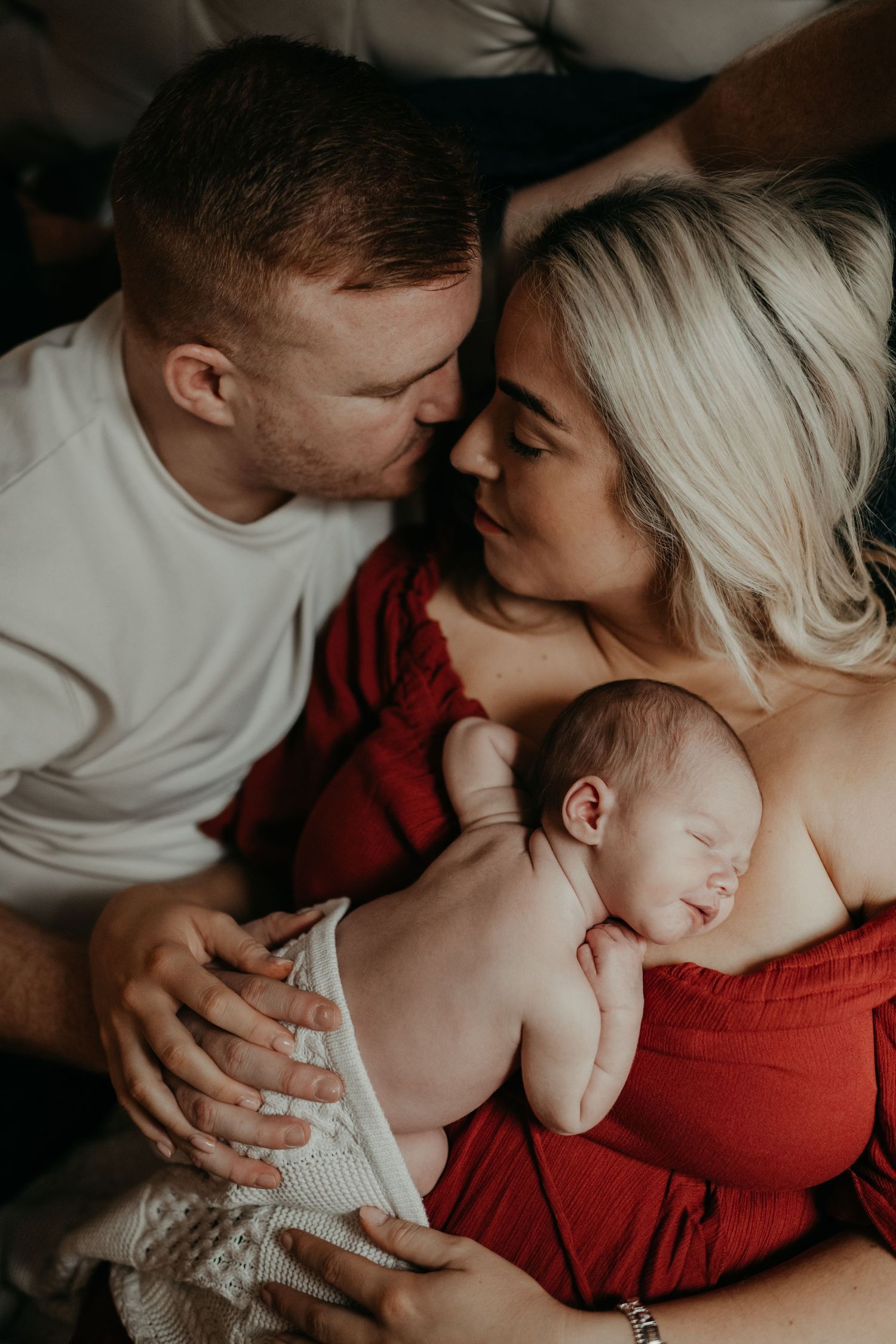 A couple lying on a bed, smiling at each other while holding their sleeping newborn baby.