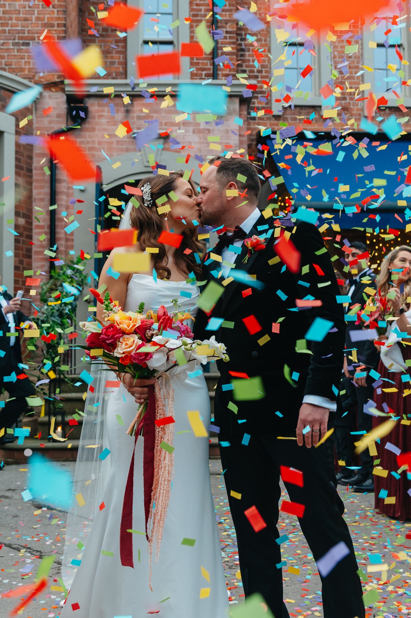 A wedding couple embraces as colorful confetti rains down around them against a brick wall backdrop.