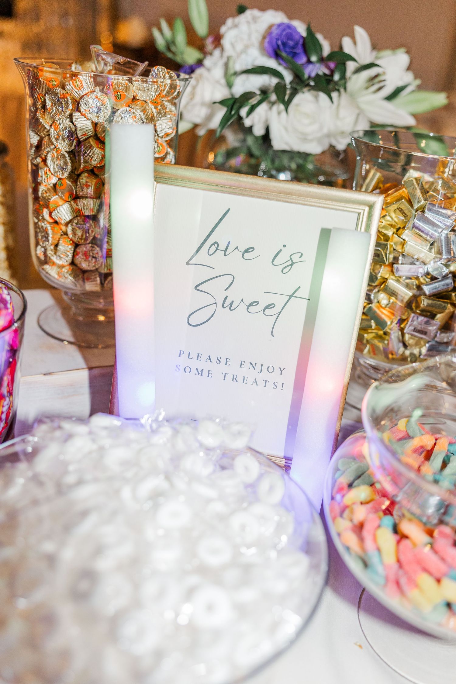 A close-up view of a wedding candy bar display with 'Love is Sweet' sign and colorful treats in glass containers.