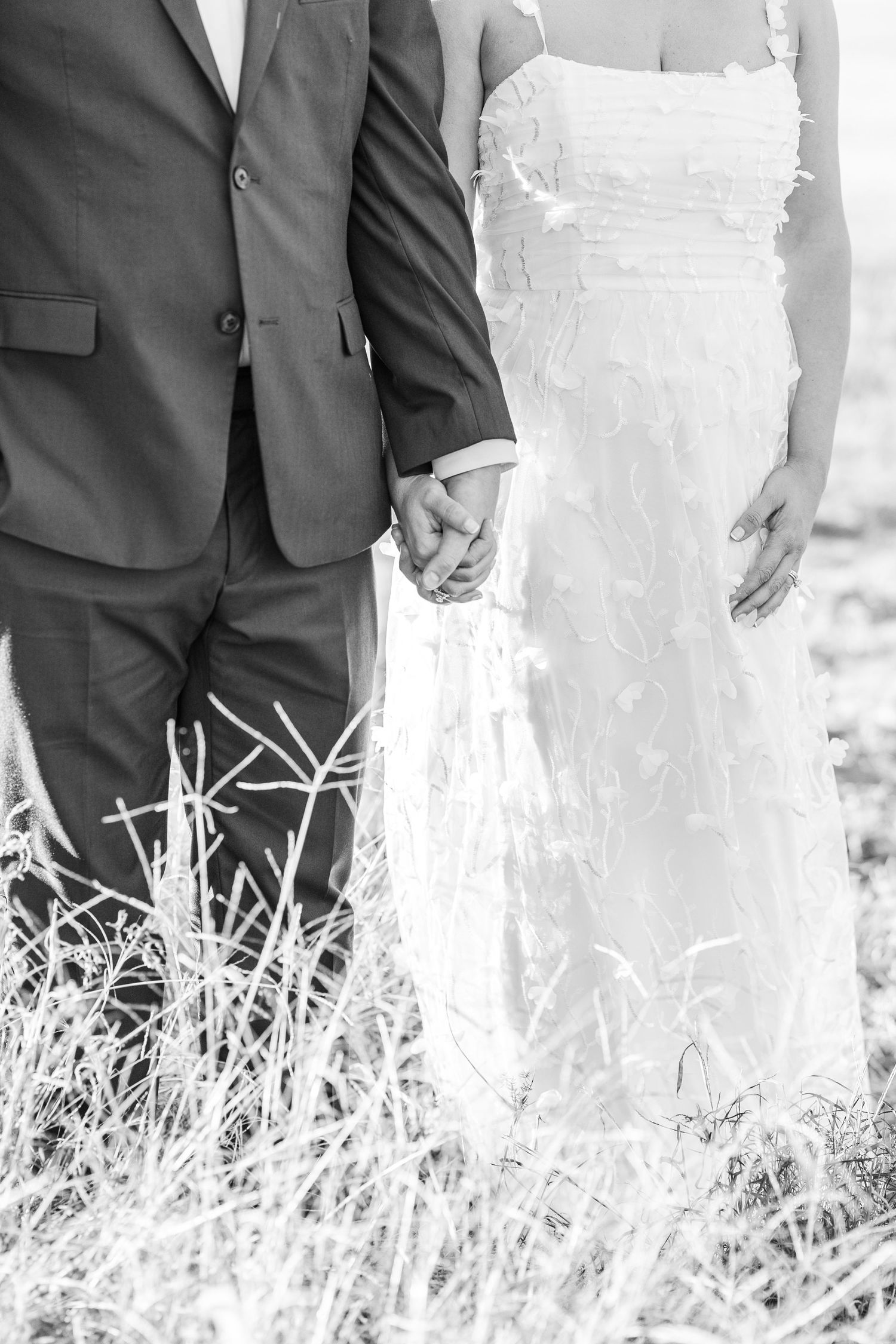 A bride and groom stand together in tall grass wearing formal wedding attire, captured in black and white.