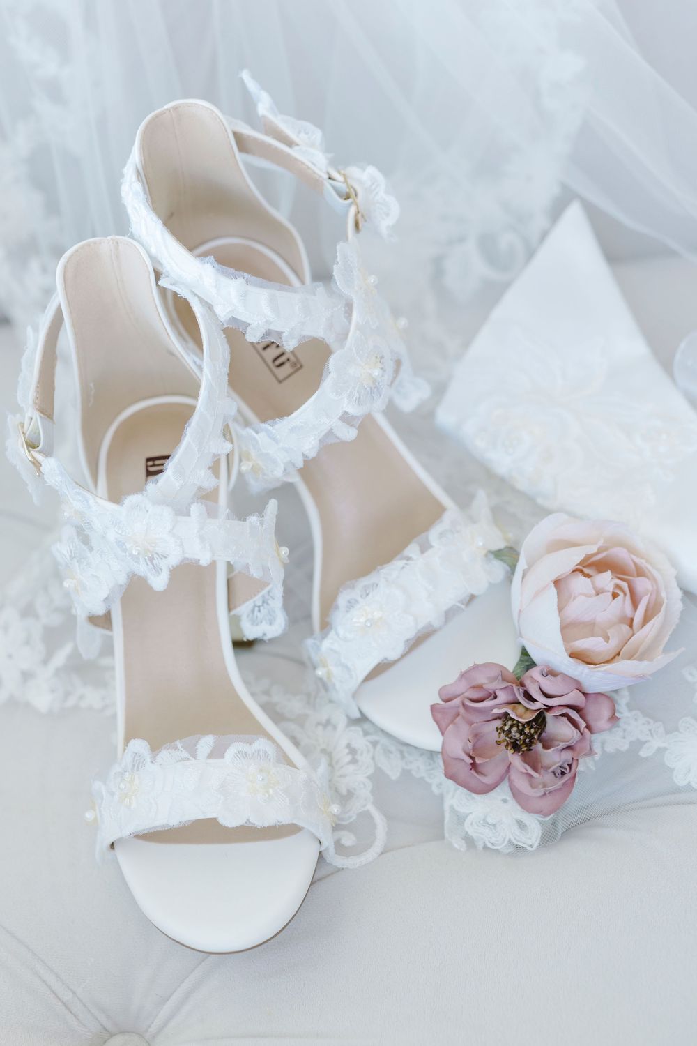 Elegant white lace bridal sandals with delicate floral details next to soft pink roses on white fabric.